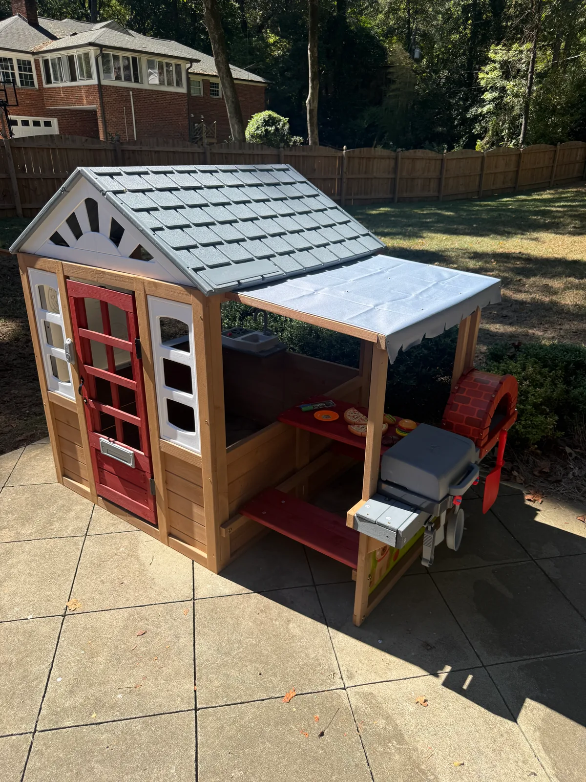 Finished wooden backyard playhouse with a red door, gray roof, canopy, and toy cooking features